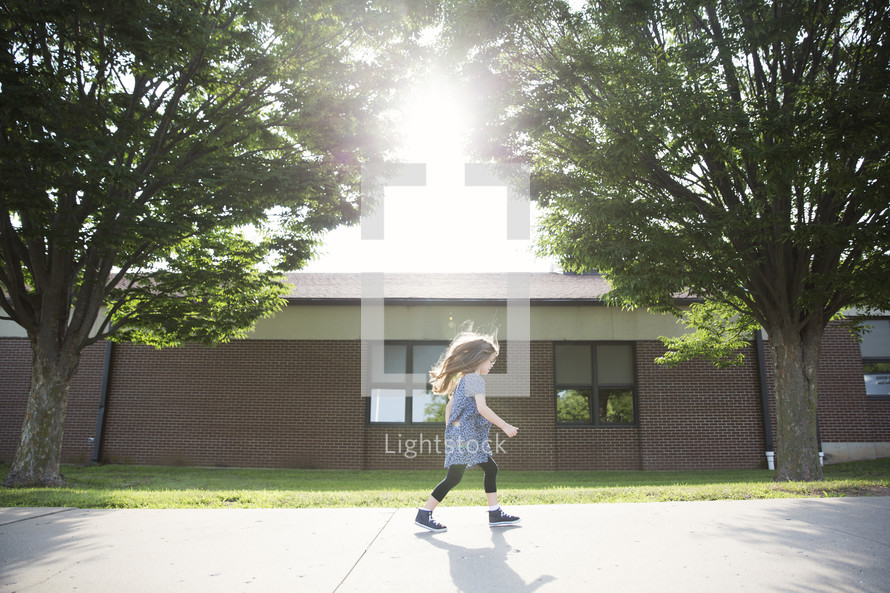 A little girl walking down the sidewalk next to school.