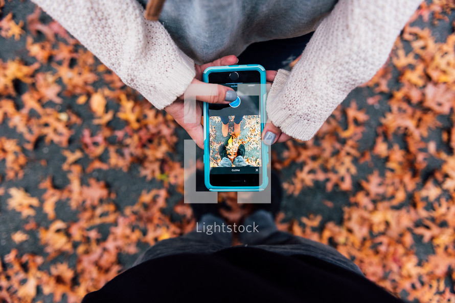 a couple with a cellphone taking a picture of their feet in fall leaves 