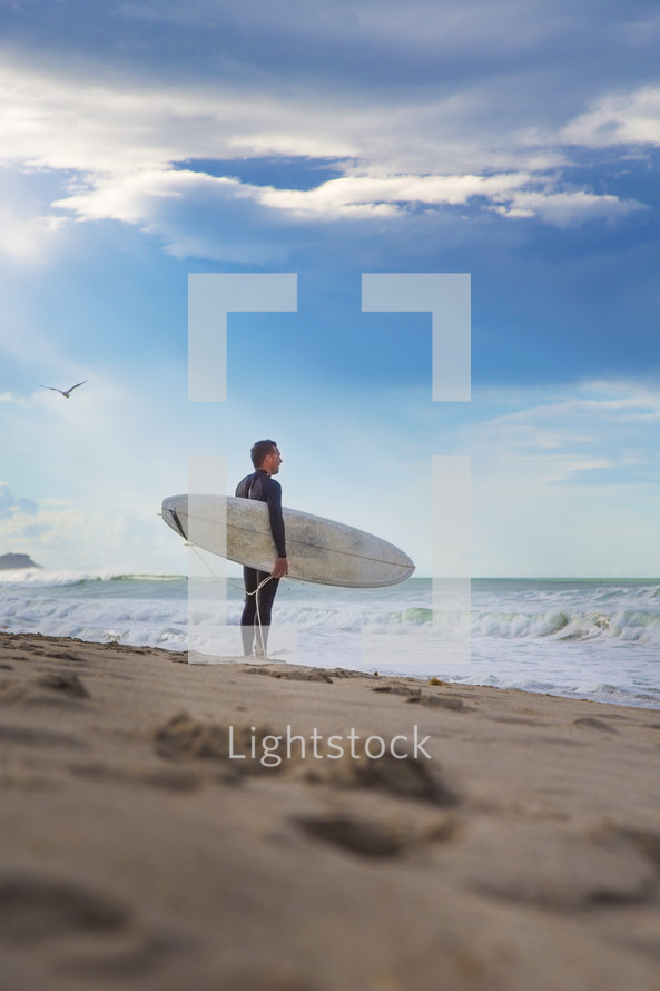 A man in a wetsuit stands on the shore carrying a surfboard.