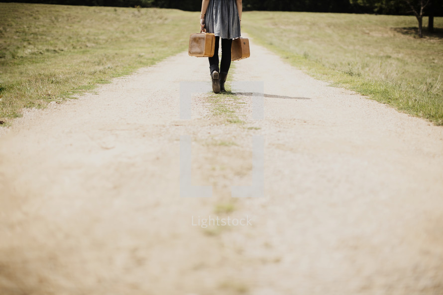 woman walking down a dirt road carrying luggage 
