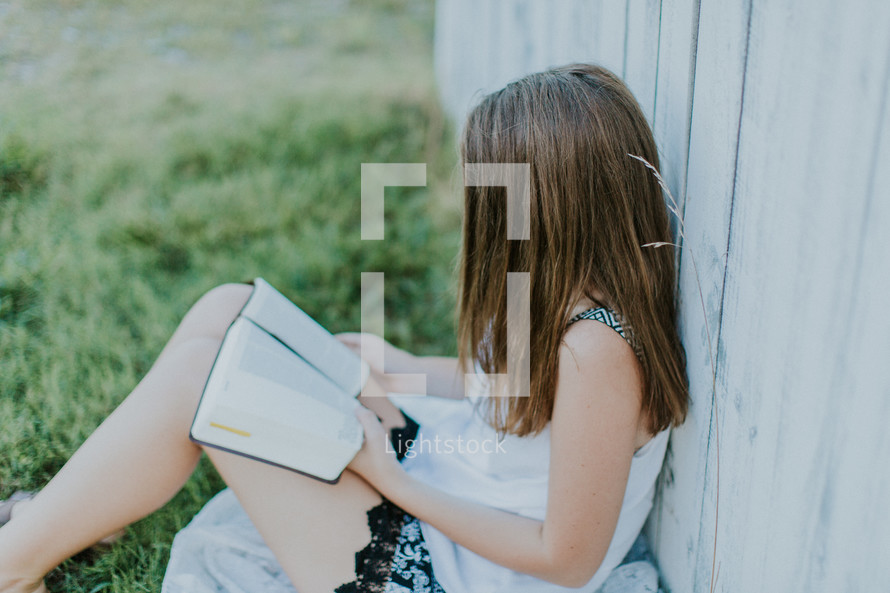 teen girl reading a Bible leaning against a fence 