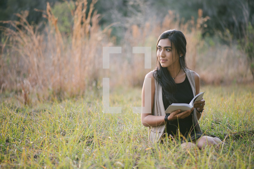 A young woman sitting outside in the grass and reading.