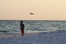 a boy watching an airplane fly over the ocean 