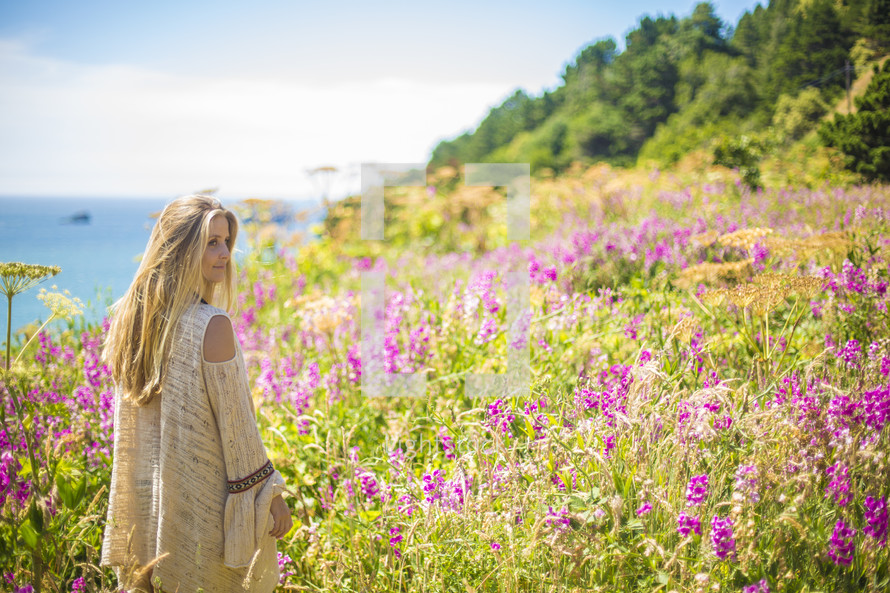 A blonde woman in a field of flowers next to the ocean.
