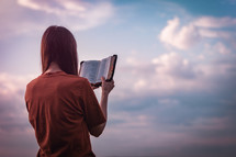 Girl reading the Bible with the sky in the background, beautiful pale color palette