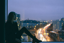 a woman sitting in a window sill looking out at a city at night 