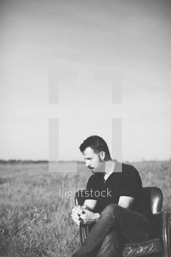 a man sitting outdoors in a leather chair in a field praying 