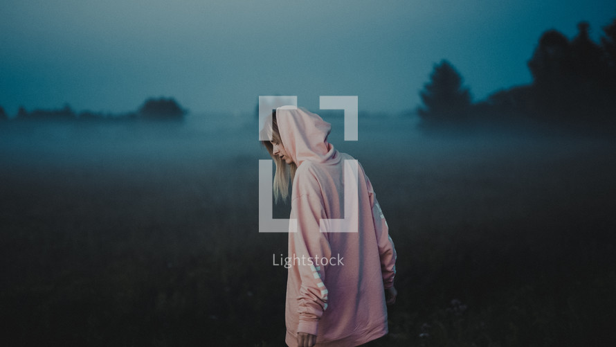 a young woman in a hoodie walking in a foggy field of tall grasses 