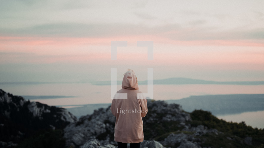 a woman in a hoodie looking out at the ocean view from a mountaintop 