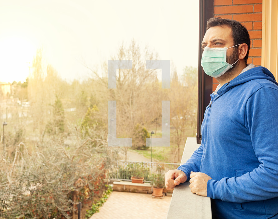 caucasian man with mask looking out on the terrace of the house during the quarantine due to the pandemic of covid19 coronavirus.