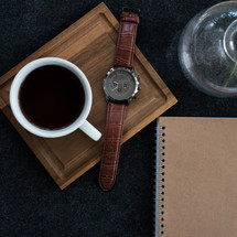 A cup of coffee and a wristwatch on a wooden board next to a vase and a spiral notebook