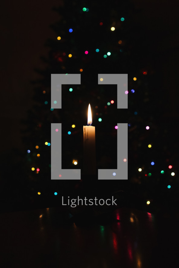 A single candle with flame in front of bokeh colorful Christmas lights on a tree