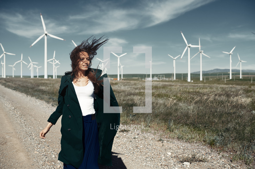 a woman walking on a dirt road beside of a wind farm 