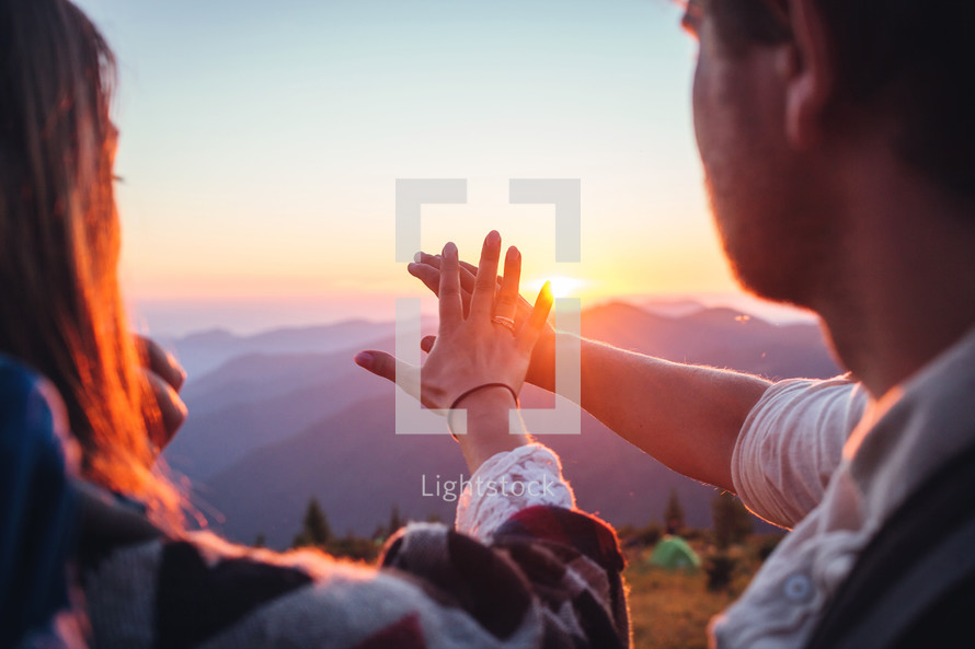 couple holding hands on a mountaintop reaching out towards the sun 