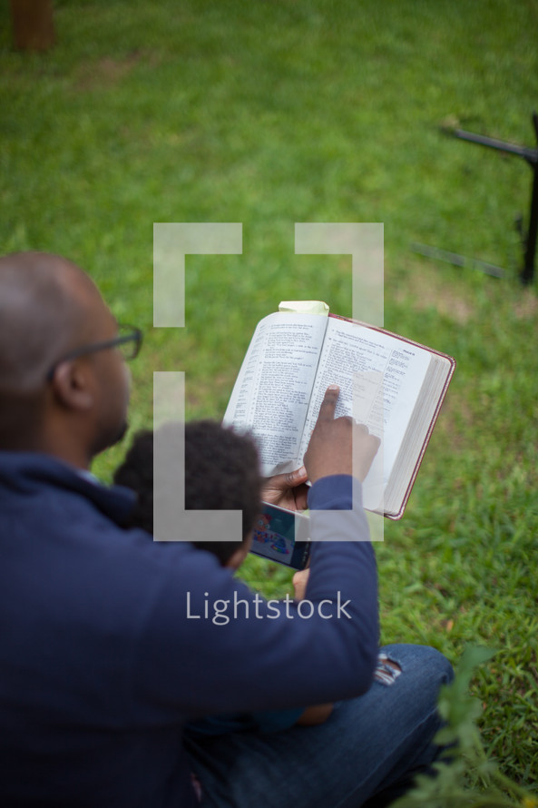A father reading the Bible with his young son in the back yard.