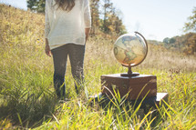 woman standing outdoors in a field of tall grass with a globe and luggage 