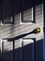 The sunlight and shadows cascade against a white wooden door that is cracked open with a brass doorknob reflecting light showing an open door or a door ready to be closed. 