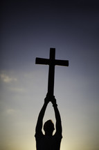 Silhouette of man holding cross over head in the air at dusk.
