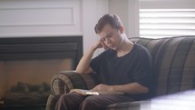a young man sits on a couch to read a Bible 