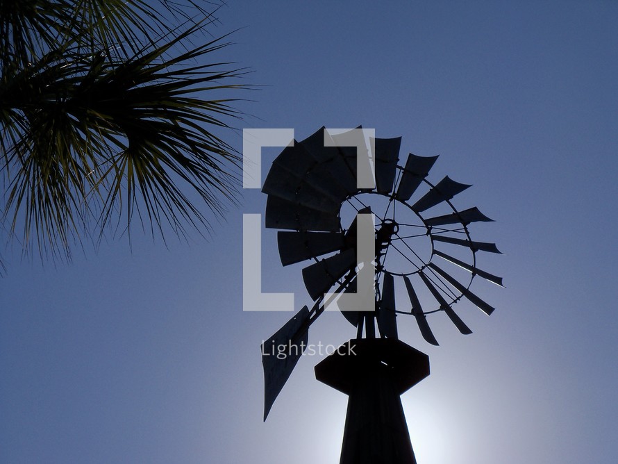 A silhouette of a wind mill standing against the sun and a clear blue sky at sunset next to palm trees on a clear, breezy and cool day. 