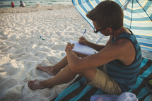 man writing in a journal on the beach under an umbrella 