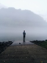 man standing on a dock near a foggy lake 