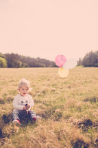 Child sitting in a field of grass holding balloons.