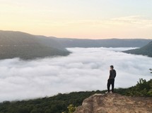 man standing on a mountaintop looking down at a foggy valley below 