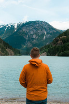 a man in an orange jacket standing by a mountain lake 