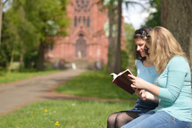 Young women smiling while reading in the bible together outside on a sunny day with a cathedral in the background. 
