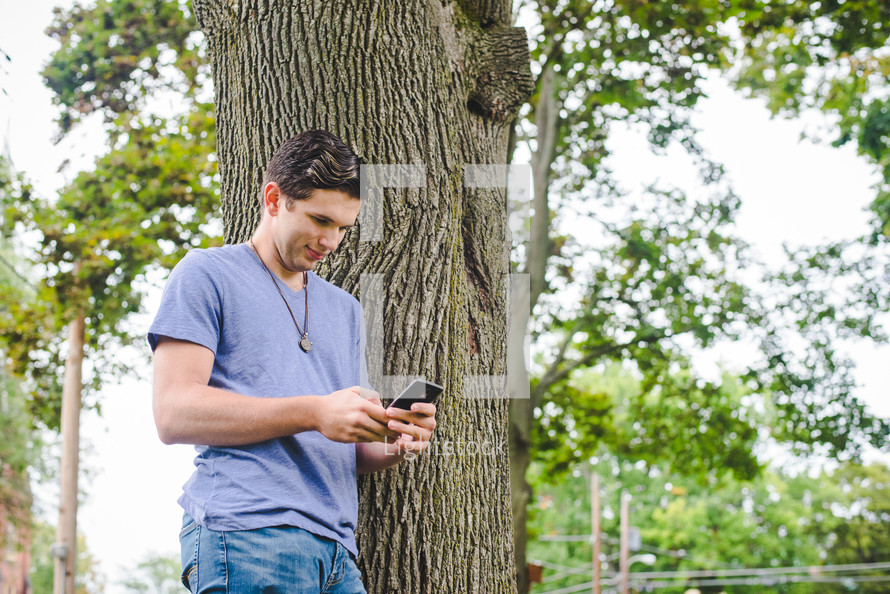 A young man standing next to a tree texting on his cellphone. 