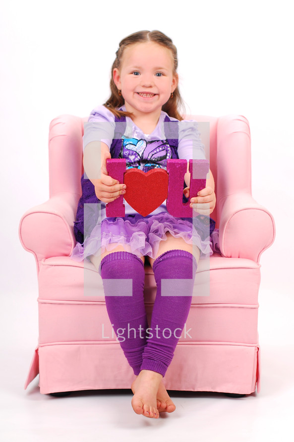 Young girl sitting in pink chair holding "I love you" sign.