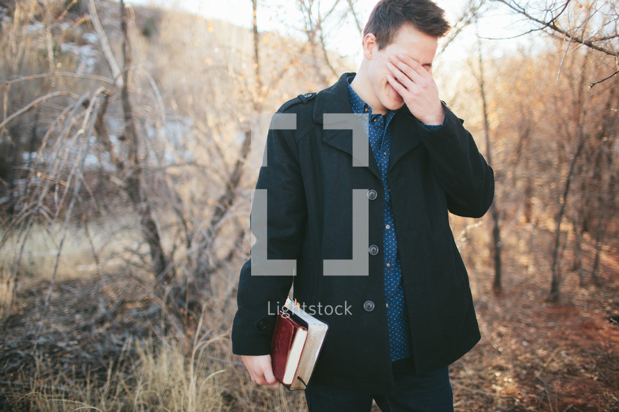 man hiding his face with his hands carrying a Bible outdoors 