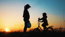 Childhood bike concept. Mother teaching son to ride bicycle. Happy cute boy learn to riding a bike in park at sunset time. Young mom teaching son to ride bike first time on countryside rural road.
