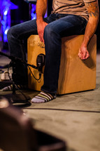 Man in jeans and socks with tattoo on arm sitting and beating on a Cajon wooden box drum on a lighted stage.