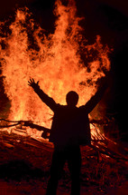 man standing with his hands raised in front of a bonfire 