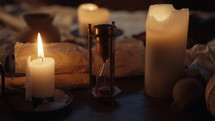 Static shot of a desk covered with open scrolls, burning candles, and an hourglass with the sand running down.