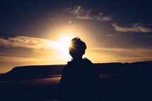 a sunburst behind a silhouette of a boy standing outdoors on a shore 