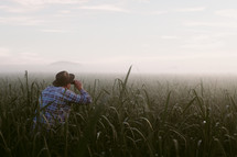 a man with binoculars standing in a field of tall green grass at dawn 