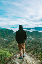 person standing at the edge looking out at green mountaintops 