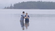 A man baptizes a young woman in the ocean with island and mountains in background.