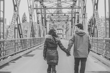 a couple holding hands walking across a bridge 