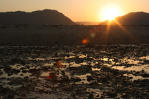 sunburst behind mountains along a shore with wet sand 