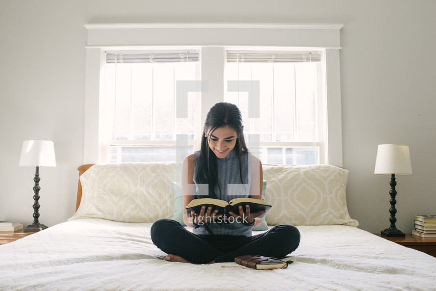 A smiling teen girl reading the Bible while sitting on her bed.