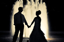 A bride and groom stand in front of a fountain.