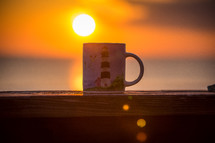 coffee mug with a lighthouse on a banister, and sun a sunrise 