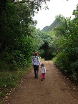 A woman and little girl walking hand-in-hand down a dirt road.