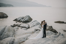 bride and groom standing on rocks along a shore 