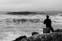 man with a fishing pole standing on rocks on a shore 