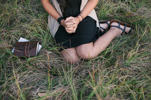 A young girl sitting in grass with hands clasped.